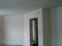 Wnętrze pomieszczenia po nałożeniu gładzi gipsowej, widoczne świeżo wykończone ściany w jasnym odcieniu, sufit z wystającymi przewodami elektrycznymi, otwór drzwiowy z widocznymi śladami po pracach...